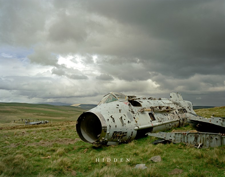 The ruined wreck of an aircraft in a field with mountains and greay clouds in the behind